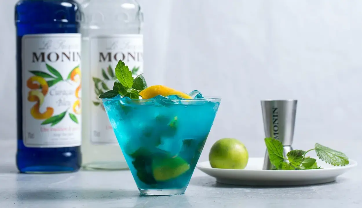 Orzeźwiający drink z syropem blue curacao smak, ozdobiony miętą i cytryną. Obok butelki syropu Monin.