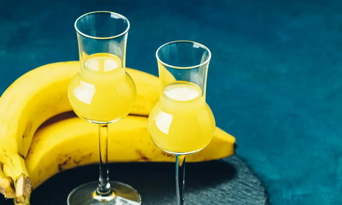 Dwa kieliszki z żółtym likierem bananowym, obok kiść bananów na ciemnoniebieskim tle.