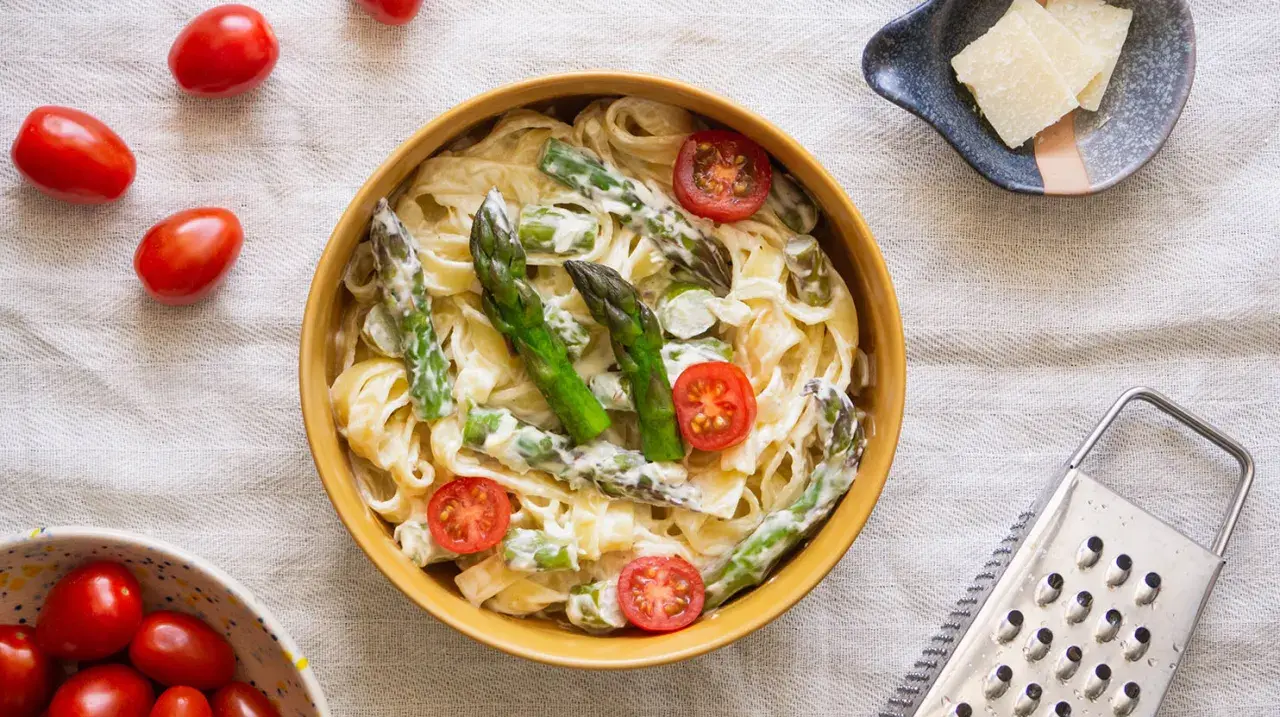 Pyszny makaron ze szparagami w kremowym sosie, ozdobiony pomidorkami koktajlowymi. Obok miseczka z parmezanem i tarką.
