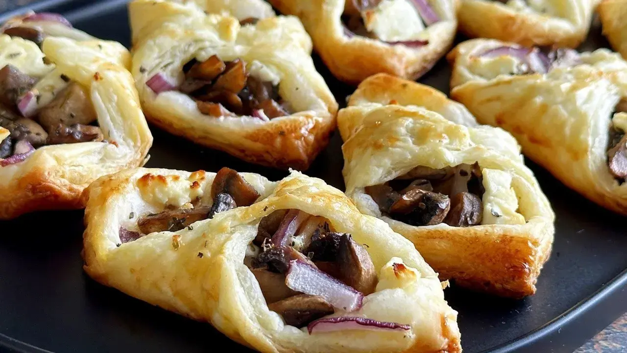 Chrupiące ciasto francuskie z pieczarkami, serem i czerwoną cebulą, ułożone na czarnej tacy.