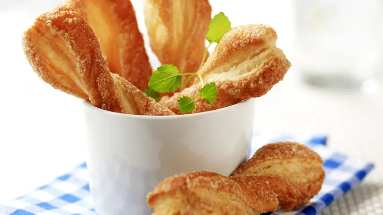 Chrupiące paluchy z ciasta francuskiego posypane cukrem, podane w białym naczyniu na niebiesko-białej kratce.