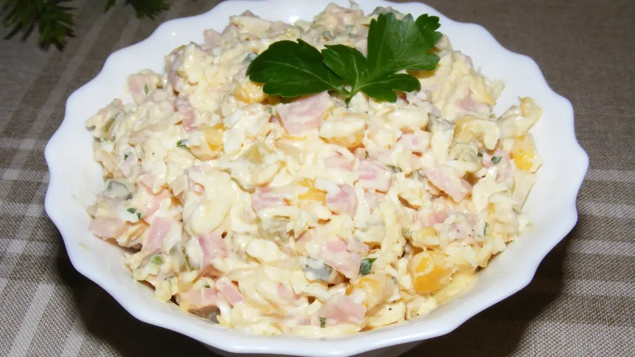 Pyszna sałatka z szynką, jajkiem, kukurydzą i ogórkiem, udekorowana natką pietruszki. Idealna na każdą okazję.