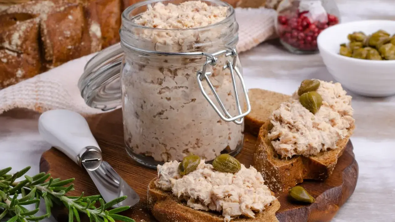 Słoik pełen pysznej pasty z tuńczyka, gotowej do rozsmarowania na chlebie. Na desce kromki z pastą i kaparami, obok gałązka rozmarynu.