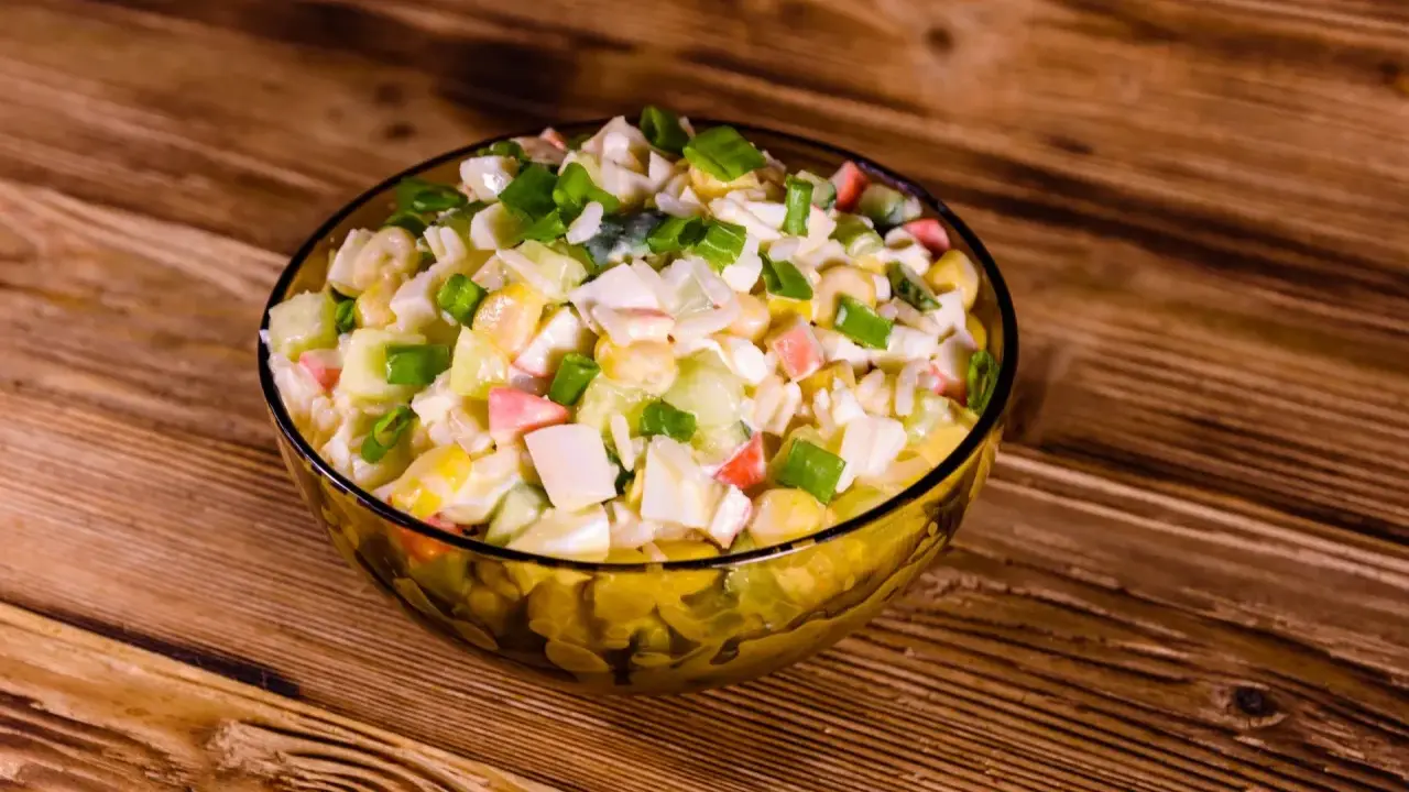 Pyszna sałatka z surimi, z kukurydzą, jajkiem i szczypiorkiem, podana w szklanej misce na drewnianym blacie.