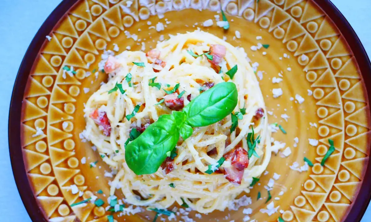 Pyszne spaghetti carbonara ze śmietaną, posypane parmezanem i natką pietruszki, ozdobione listkiem bazylii.