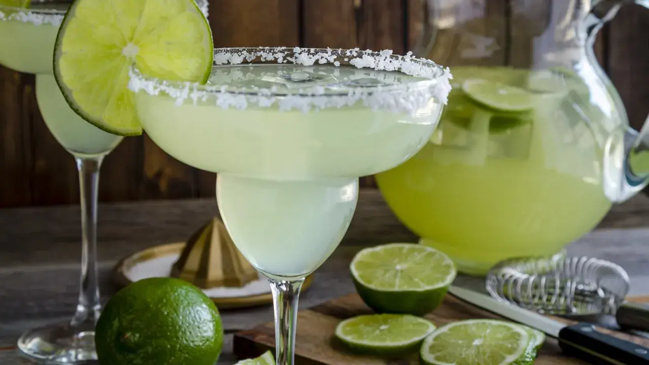 Orzeźwiająca margarita z solą na brzegu kieliszka, ozdobiona plasterkiem limonki. Obok dzbanek z drinkiem i świeże limonki.