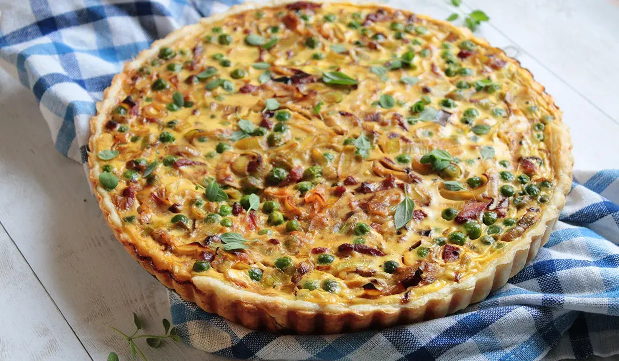 Pyszna tarta z porem i boczkiem, udekorowana groszkiem i ziołami, na niebiesko-białej kratce.