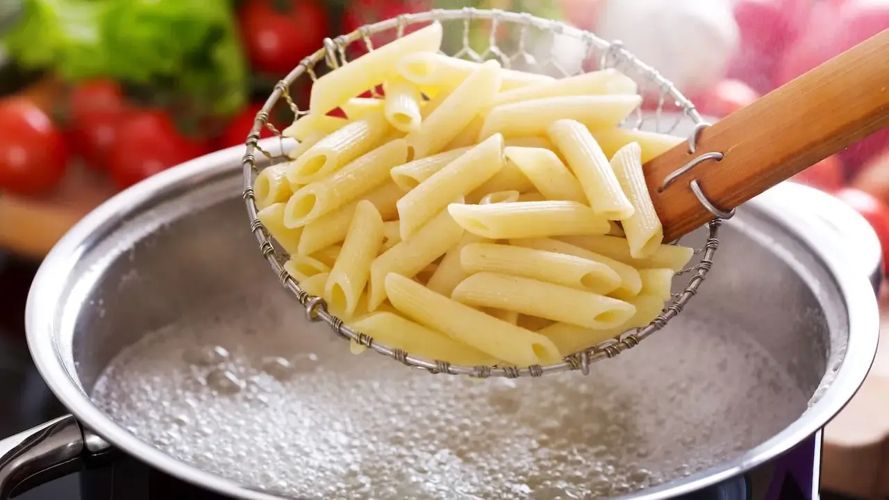 Penne makaron w cedzaku nad garnkiem z wrzącą wodą. Czas, ile się gotuje makaron, zależy od jego rodzaju.