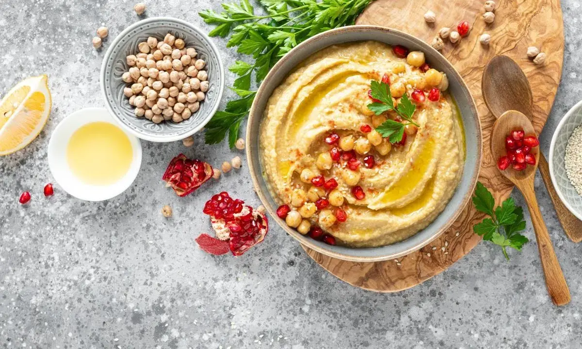 Pyszny hummus z ciecierzycy, udekorowany pestkami granatu i natką pietruszki, obok cytryny i oliwy.