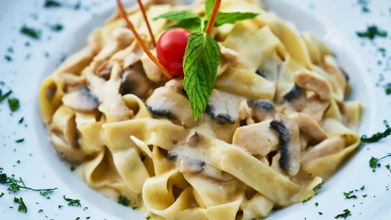 Pyszny makaron tagliatelle z kremowym sosem pieczarkowym, udekorowany pomidorkiem koktajlowym i listkiem mięty.