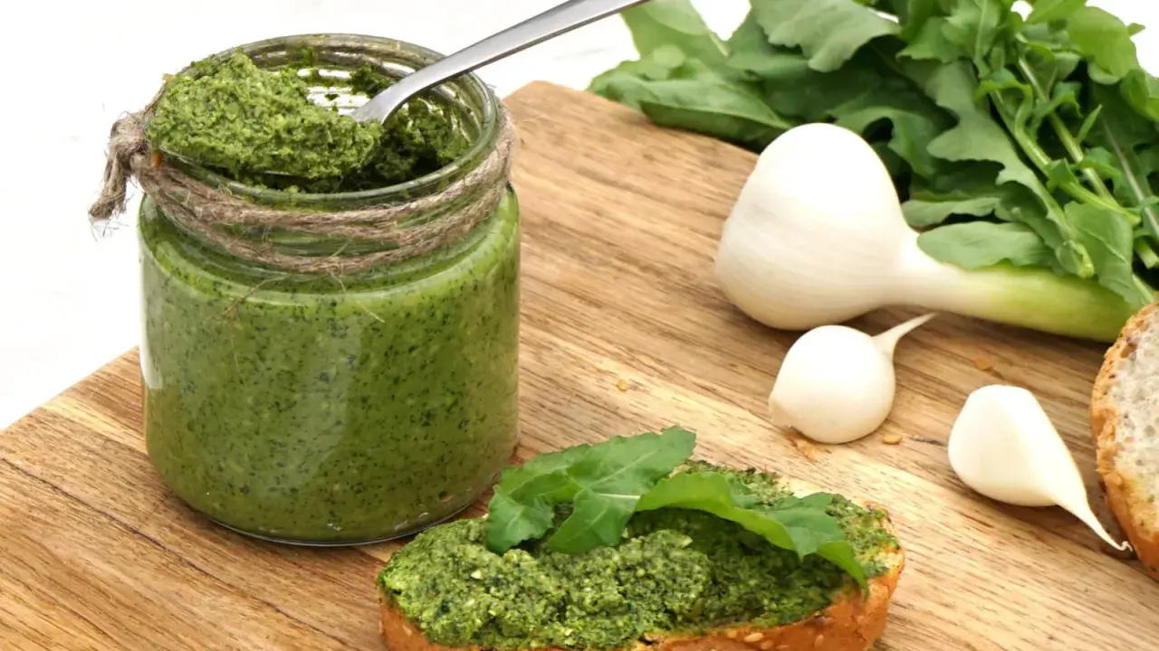 Słoik domowego pesto z rukoli, łyżka w środku, kromka chleba z pastą i świeże składniki: czosnek i rukola.