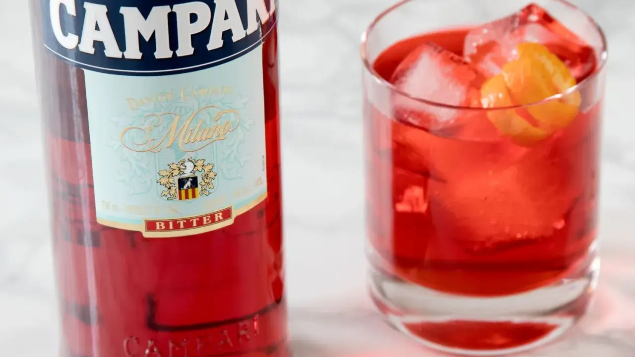 Butelka Campari i szklanka z czerwonym drinkiem, lodem i skórką pomarańczy. Idealny aperitif, który może być bazą do drinków z Aperolem.