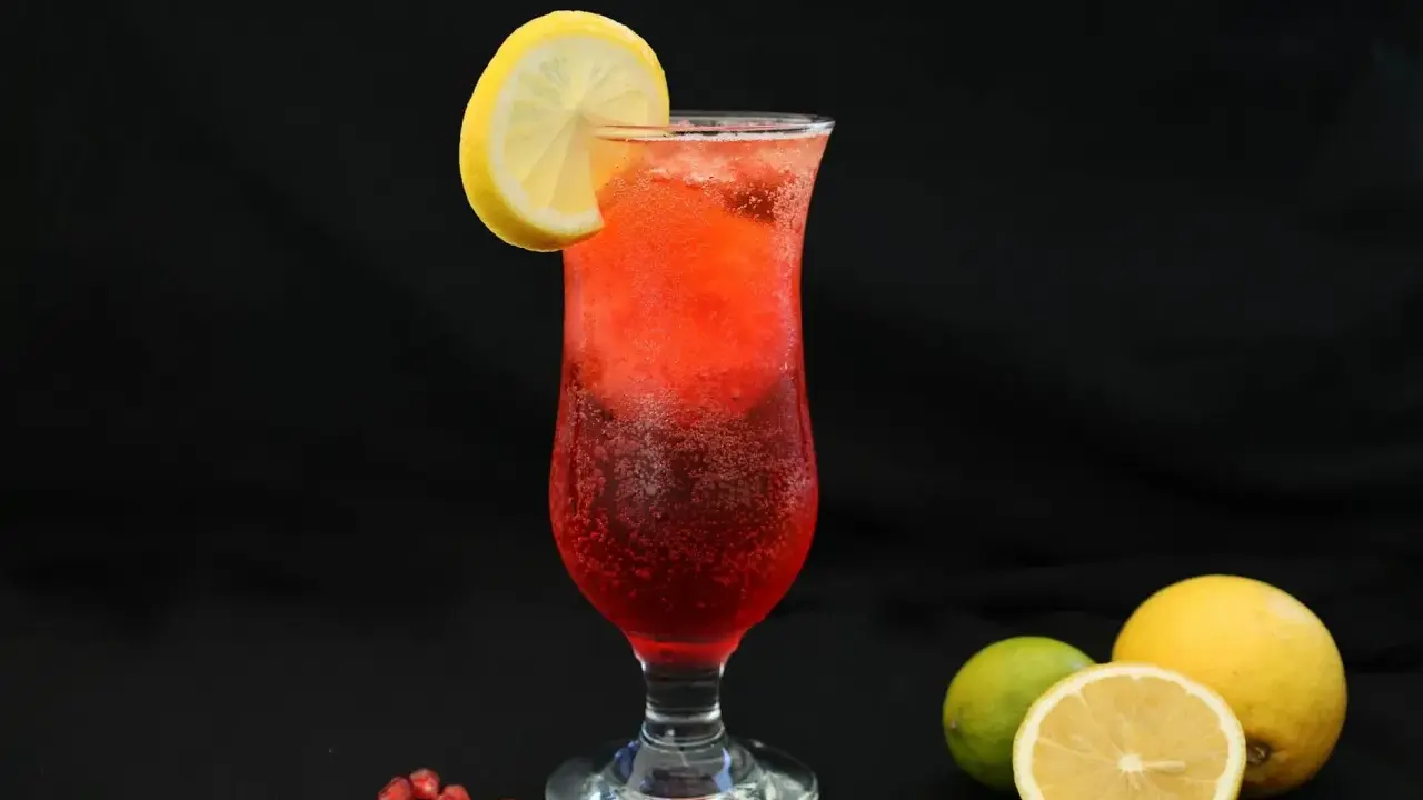 Orzeźwiający drink z granatem, ozdobiony plasterkiem cytryny. Obok leżą świeże owoce: cytryna i limonka.