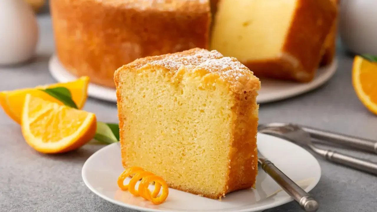 Puszysta babka pomarańczowa, oprószona cukrem pudrem, z plasterkami pomarańczy i ozdobną skórką na talerzyku.