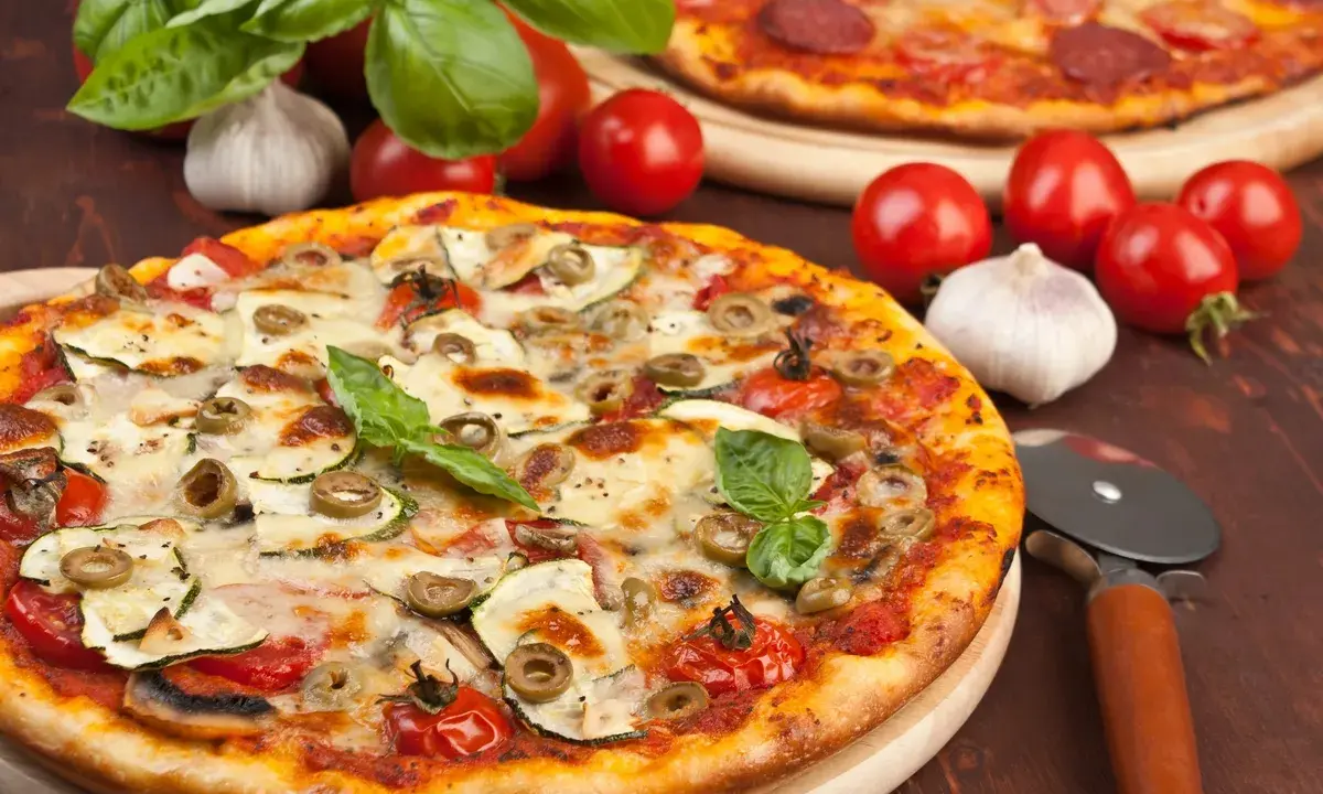 Pizza z cukinią, oliwkami i pomidorkami, obok surowe pieczarki na pizze, czosnek i bazylia.
