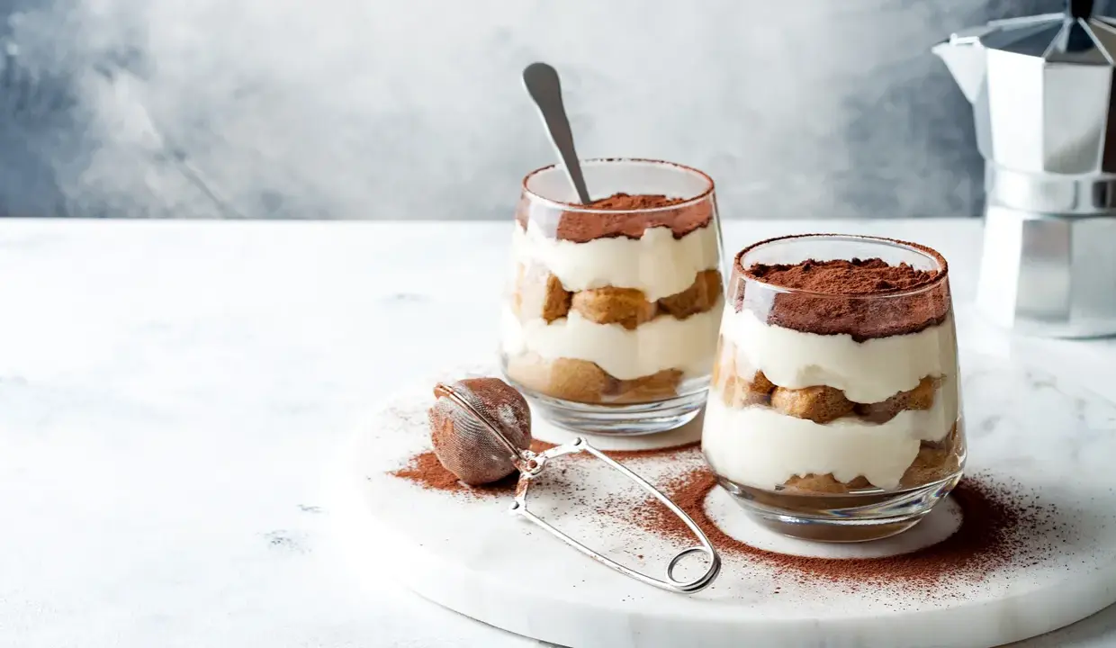 Dwa pucharki z tiramisu bez jajek, posypane kakao, na marmurowej desce.