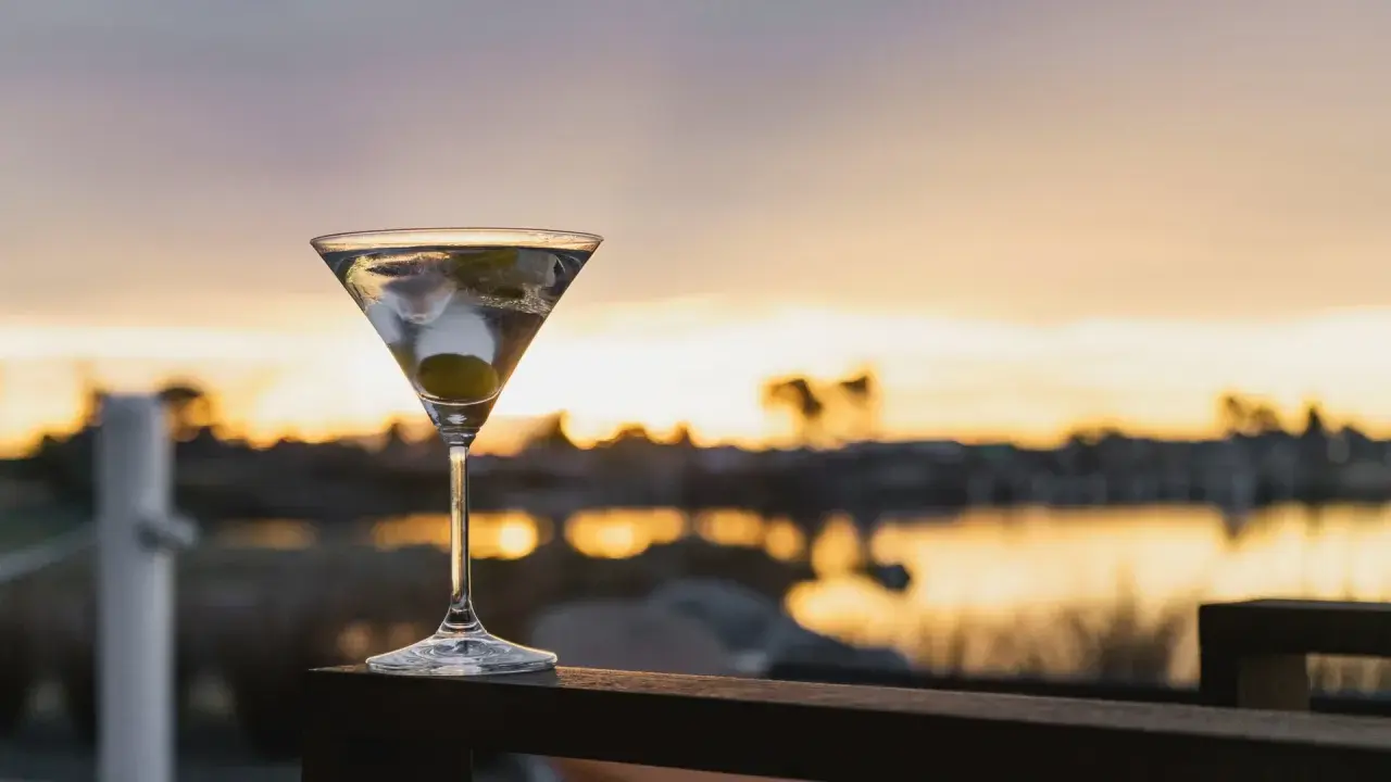 Idealny martini wstrząśnięte nie mieszane, z oliwką, na tle zachodzącego słońca nad wodą.