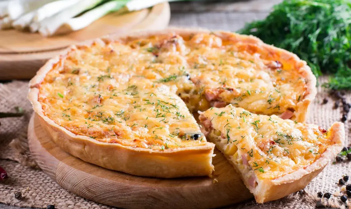 Złocista tarta z porem, posypana ziołami, z kawałkiem odciętym na desce. Obok pęczek pora i koperek.