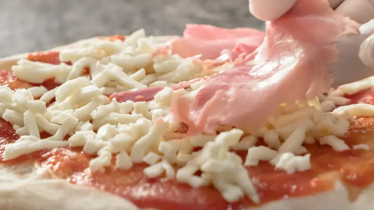 Ręce w białych rękawiczkach układają cienkie plastry szynki do pizzy na serze i sosie pomidorowym.