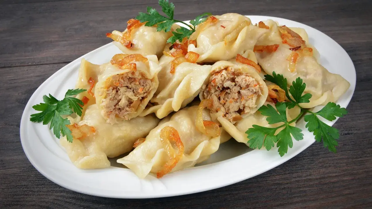 Pyszne pierogi z mięsem i kapustą, okraszone skwarkami i udekorowane natką pietruszki, na białym talerzu.