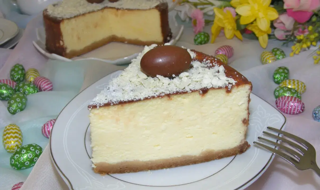 Pyszny sernik z mascarpone, udekorowany czekoladowymi jajeczkami i wiórkami białej czekolady, na tle wielkanocnych ozdób.