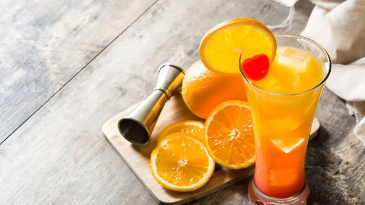 Kolorowy drink tequila sunrise z pomarańczą i wisienką, obok pomarańcze i miarka barmańska.