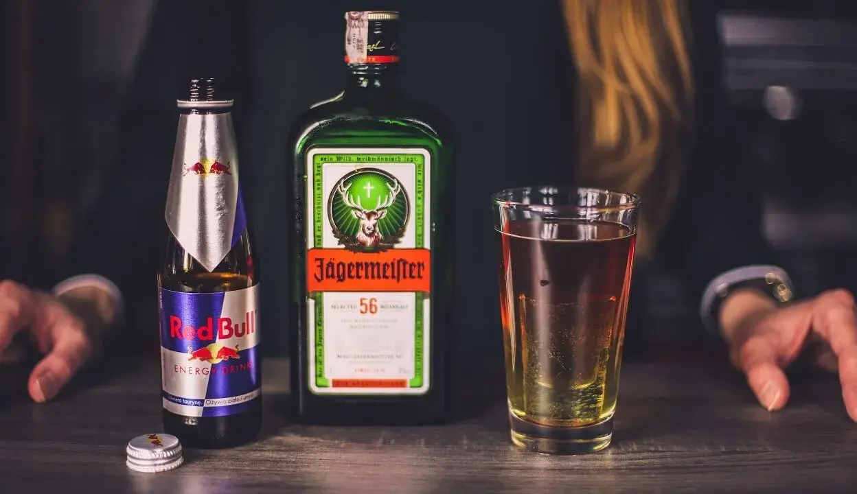 Jägermeister z czym pić? Klasycznie z Red Bullem, tworząc popularny "Jägerbomb". Na stole butelka Jägermeistera, Red Bull i szklanka z drinkiem.