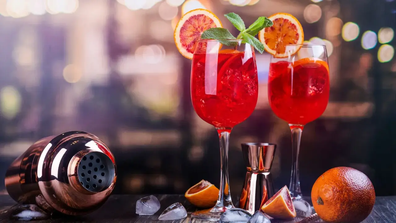 Dwa kieliszki orzeźwiającego drinka z prosecco z czymś czerwonym, plasterkami pomarańczy i miętą. Obok shaker i pomarańcze.