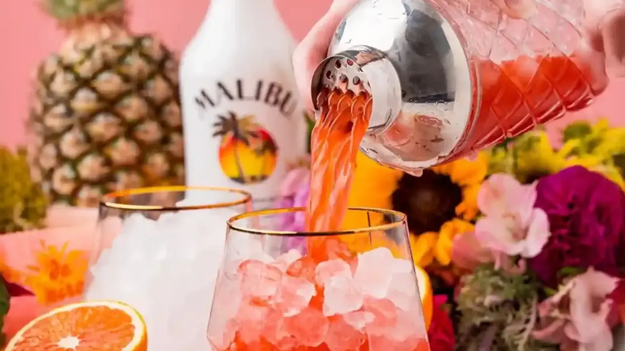 Malibu z sokiem ananasowym nalewany do szklanki z lodem. W tle ananas, butelka Malibu i kwiaty.