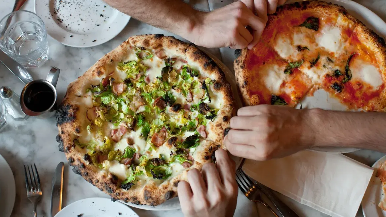 Dwie pizze na stole, jedna z brukselką i boczkiem, druga margherita. Ludzie sięgają po kawałki, pokazując, ile wyrasta ciasto na pizze.