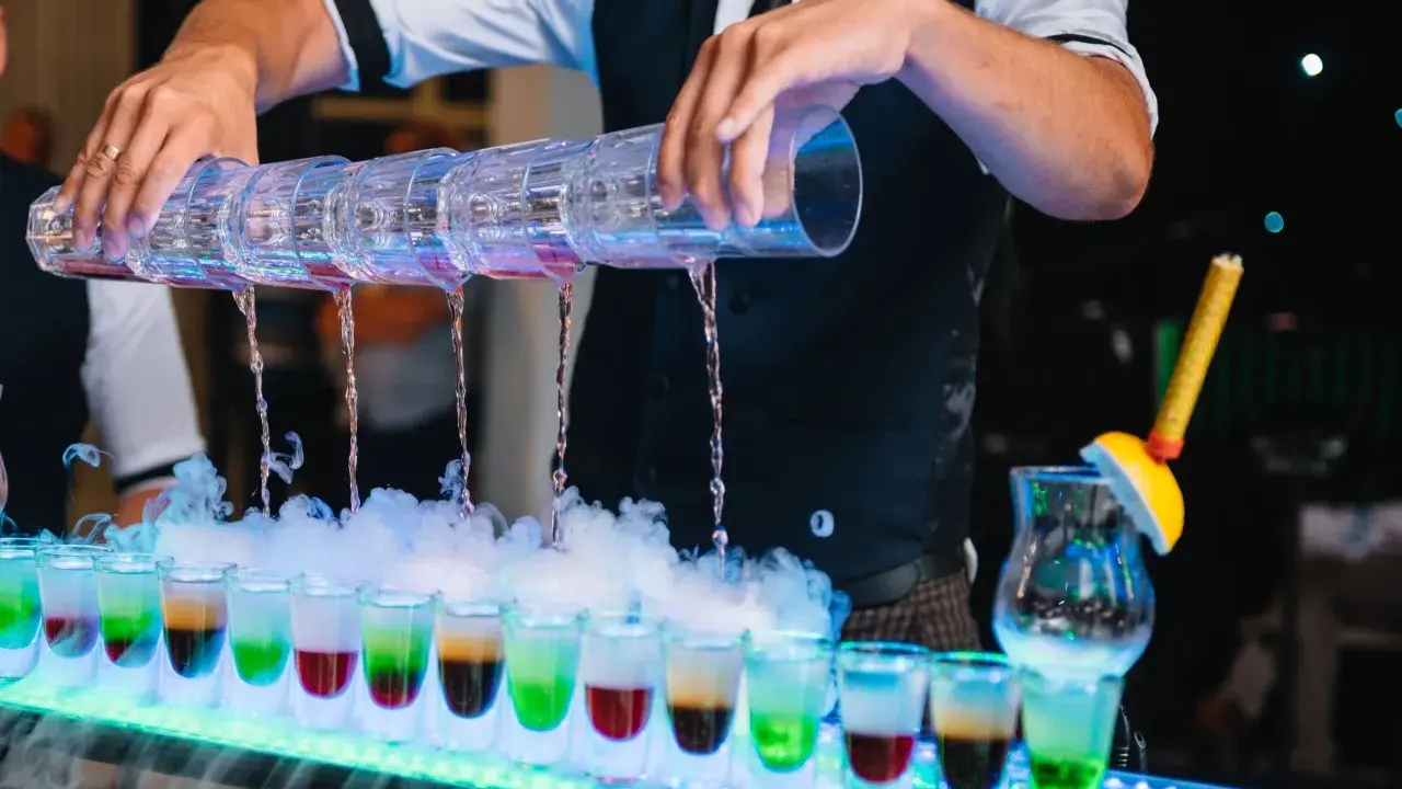 Barman nalewa kolorowe shoty z wielopoziomowego shakera. Efektowny drink bar diy z dymem i neonowym oświetleniem.