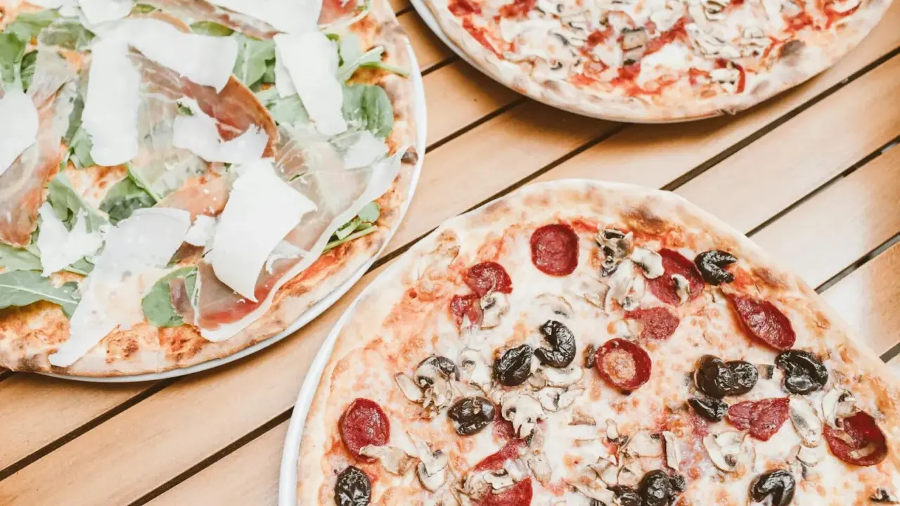 Trzy pizze na drewnianym stole. Jedna z szynką parmeńską i rukolą, druga z salami i pieczarkami, trzecia z pieczarkami. Szukasz alternatywy, jaki ser do pizzy zamiast mozzarelli?