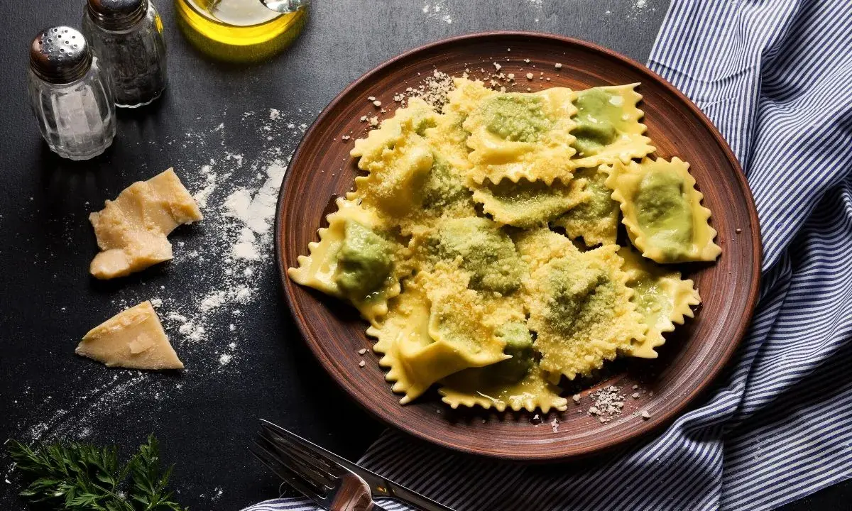 Pyszne ravioli z ricottą i szpinakiem, posypane parmezanem, na brązowym talerzu. Obok kawałki sera i przyprawy.