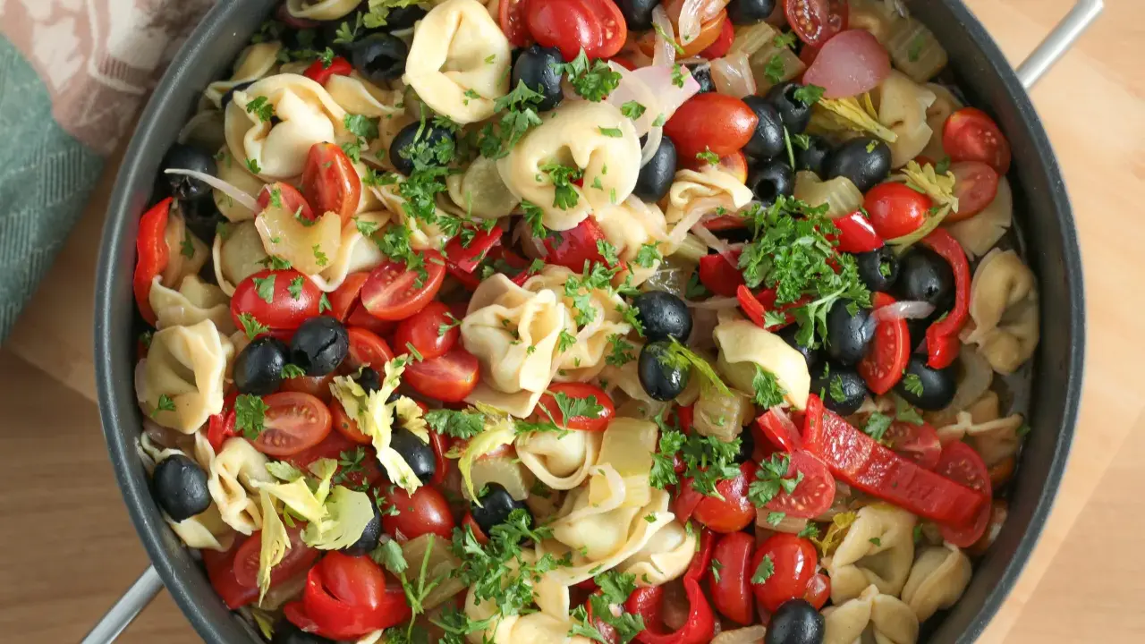 Kolorowa sałatka z tortellini, pomidorkami koktajlowymi, czarnymi oliwkami, papryką i natką pietruszki w dużej patelni.