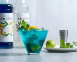 Orzeźwiający drink z syropem blue curacao smak, ozdobiony miętą i cytryną. Obok butelki syropu Monin.