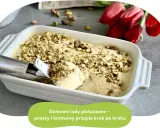 Kremowe lody pistacjowe w białej formie, posypane orzechami. Łyżka właśnie nabiera porcję tego pysznego deseru.