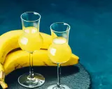 Dwa kieliszki z żółtym likierem bananowym, obok kiść bananów na ciemnoniebieskim tle.