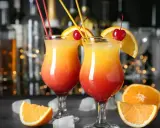 Dwa kolorowe, domowe drinki z wódką, ozdobione pomarańczą i wiśnią, z kostkami lodu i owocami w tle.