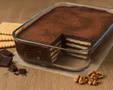 Pyszne ciasto bez pieczenia z warstwami herbatników i czekoladowego kremu, posypane kakao. Obok leżą herbatniki, kawałki czekolady i orzechy włoskie.