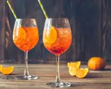 Dwa orzeźwiające Aperol Spritz z plasterkami pomarańczy i słomkami, na drewnianym stole.