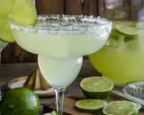 Orzeźwiająca margarita z solą na brzegu kieliszka, ozdobiona plasterkiem limonki. Obok dzbanek z drinkiem i świeże limonki.