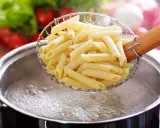 Penne makaron w cedzaku nad garnkiem z wrzącą wodą. Czas, ile się gotuje makaron, zależy od jego rodzaju.