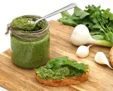 Słoik domowego pesto z rukoli, łyżka w środku, kromka chleba z pastą i świeże składniki: czosnek i rukola.