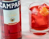 Butelka Campari i szklanka z czerwonym drinkiem, lodem i skórką pomarańczy. Idealny aperitif, który może być bazą do drinków z Aperolem.