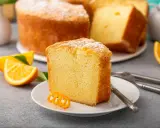 Puszysta babka pomarańczowa, oprószona cukrem pudrem, z plasterkami pomarańczy i ozdobną skórką na talerzyku.