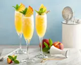 Trzy orzeźwiające drinki mimoza w kieliszkach flute, ozdobione brzoskwinią i miętą, na marmurowym blacie.