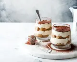 Dwa pucharki z tiramisu bez jajek, posypane kakao, na marmurowej desce.