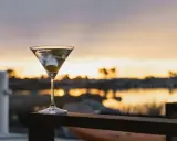 Idealny martini wstrząśnięte nie mieszane, z oliwką, na tle zachodzącego słońca nad wodą.