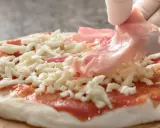Ręce w białych rękawiczkach układają cienkie plastry szynki do pizzy na serze i sosie pomidorowym.