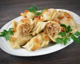 Pyszne pierogi z mięsem i kapustą, okraszone skwarkami i udekorowane natką pietruszki, na białym talerzu.